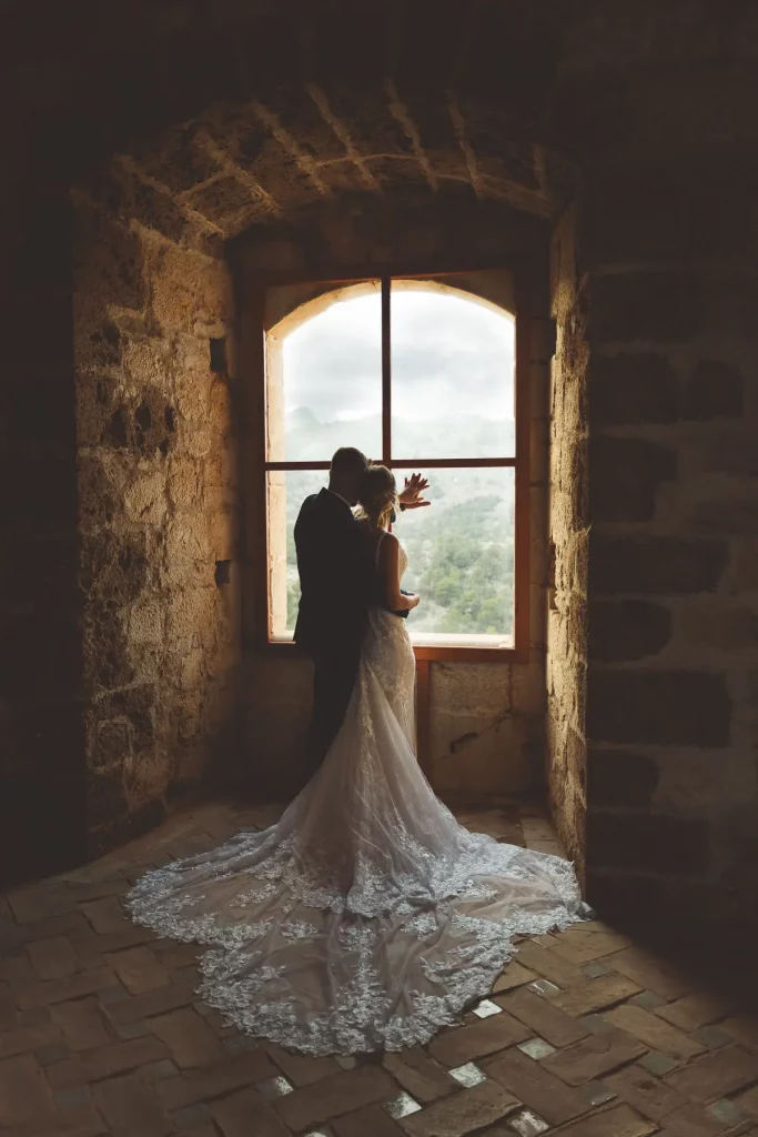 Foto creativa de unos novios en su postboda en un castillo mirando por la ventana en Almería, con la luz del atardecer entrando por ella